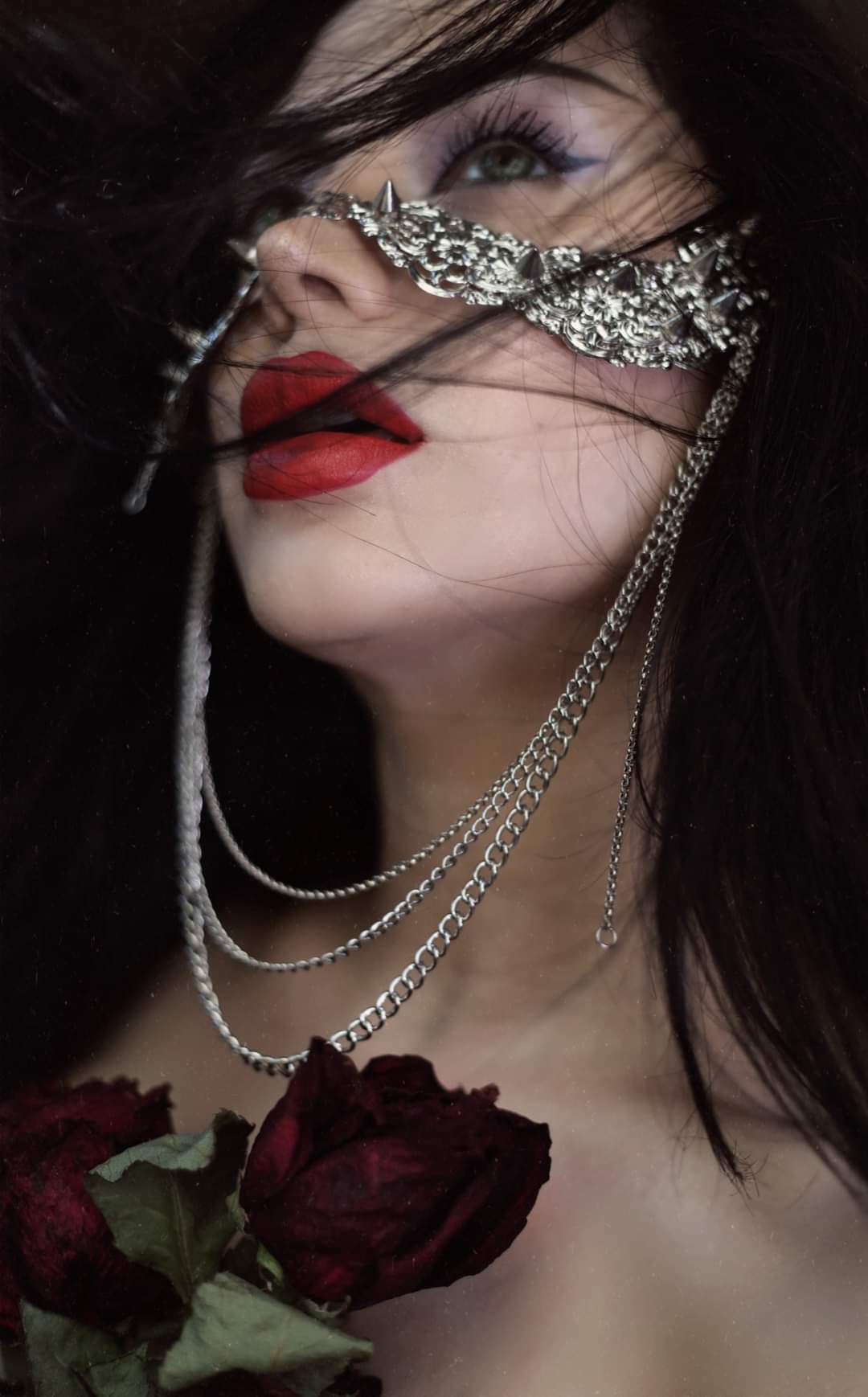 A model from Myril Jewels exudes a dark-avantgarde aura, adorned with a neo-goth studded nose mask featuring hanging chains, blurring the lines between gothic-chic and whimsigoth styles. The mask, a statement piece for Halloween or festival wear, showcases intricate craftsmanship, perfect for alternative style lovers. It pairs strikingly with the model's vivid red lips and dark, flowing hair, capturing a witchcore essence suitable for both everyday wear and rave party fashion.