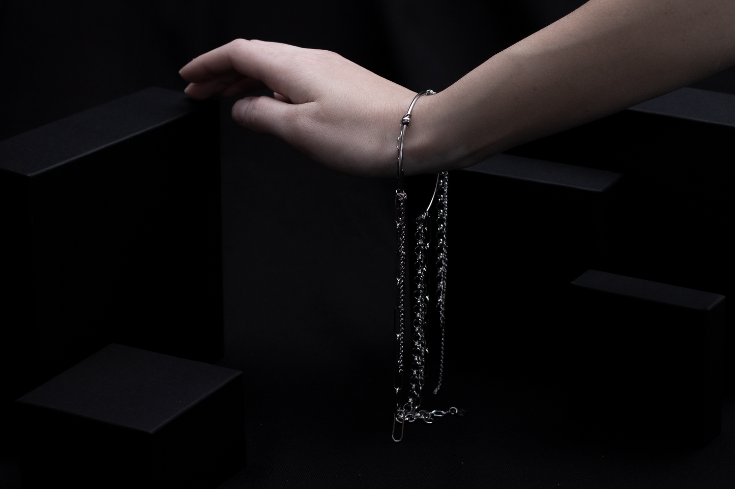 A wrist adorned with a Myril Jewels rigid bracelet is elegantly positioned against a dark background, with multiple chains draping gracefully from it. The bracelet’s design exudes gothic allure, making it an impeccable choice for those embracing dark-avantgarde fashion, whether it’s for daily sophistication or a special event.