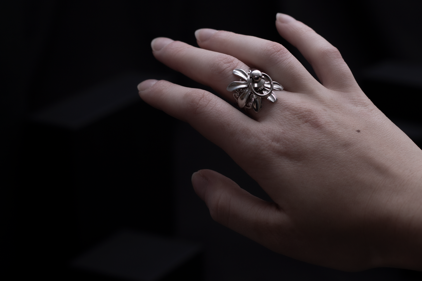 A solitary hand is gracefully adorned with a Myril Jewels ring, capturing the essence of dark-avantgarde elegance. The silver ring, featuring a distinctive floral motif with a gothic twist, sits prominently against the contrasting dark backdrop, designed for those with a passion for gothic and alternative styles. This piece is a testament to Myril Jewels' commitment to crafting unique, handmade jewelry that embodies an avant-garde and sophisticated Italian aesthetic.