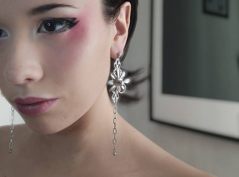 An elegant model showcases Myril Jewels' neo-gothic earring, a statement piece of jewelry for gothic and alternative style aficionados. The design features a floral motif with a cascading chain, exemplifying dark-avantgarde aesthetics. Ideal for Halloween, it complements minimal goth attire and makes a bold gift for a goth girlfriend or friend. The accessory resonates with the whimsigoth trend, perfect for everyday wear or as a standout rave party or festival jewel