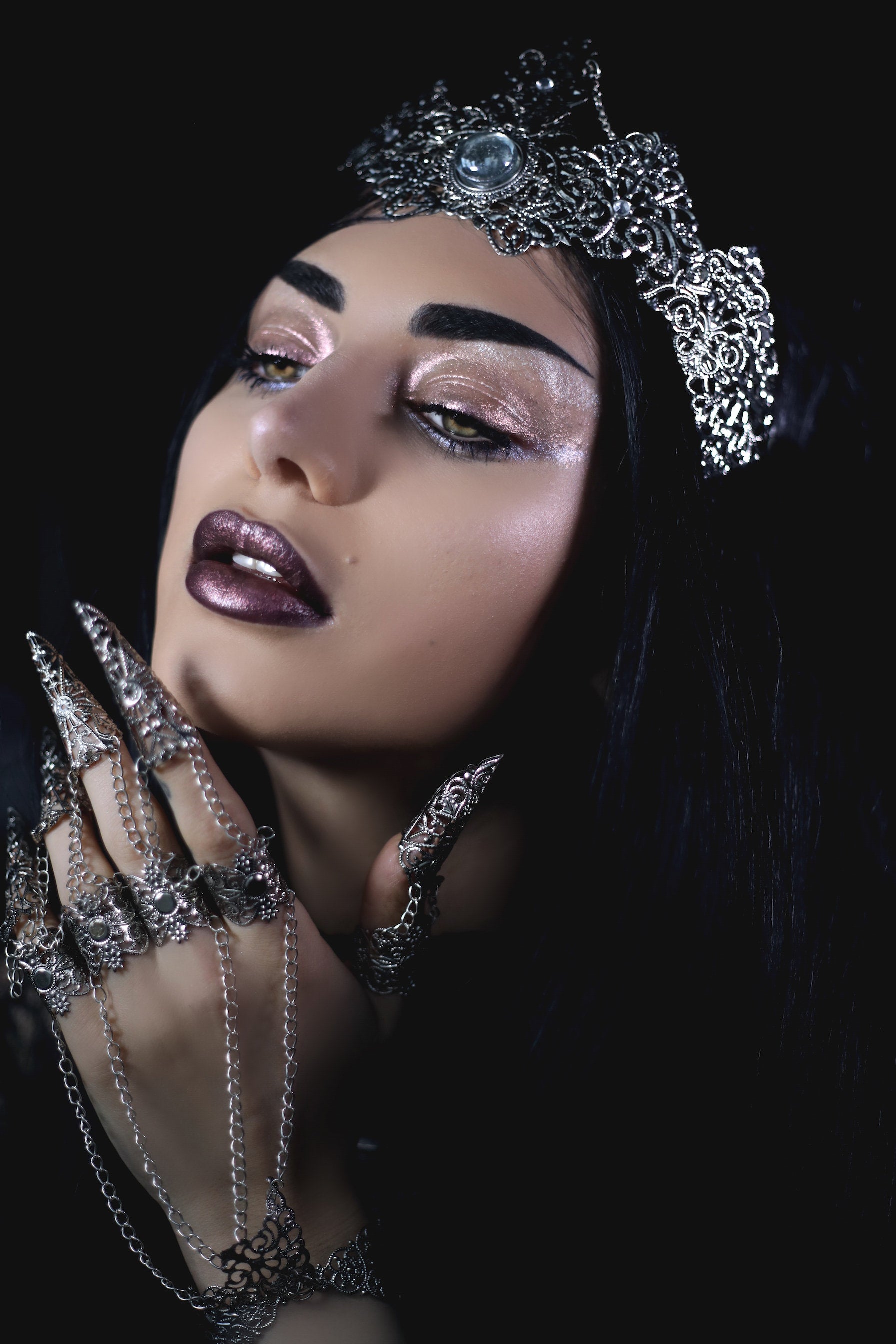 Portrait of a woman embodying gothic elegance with shimmering dark makeup and silver jewelry, including a detailed headpiece, chain-linked claw rings, and an ornate necklace, resonating with a vampiric goth aesthetic.