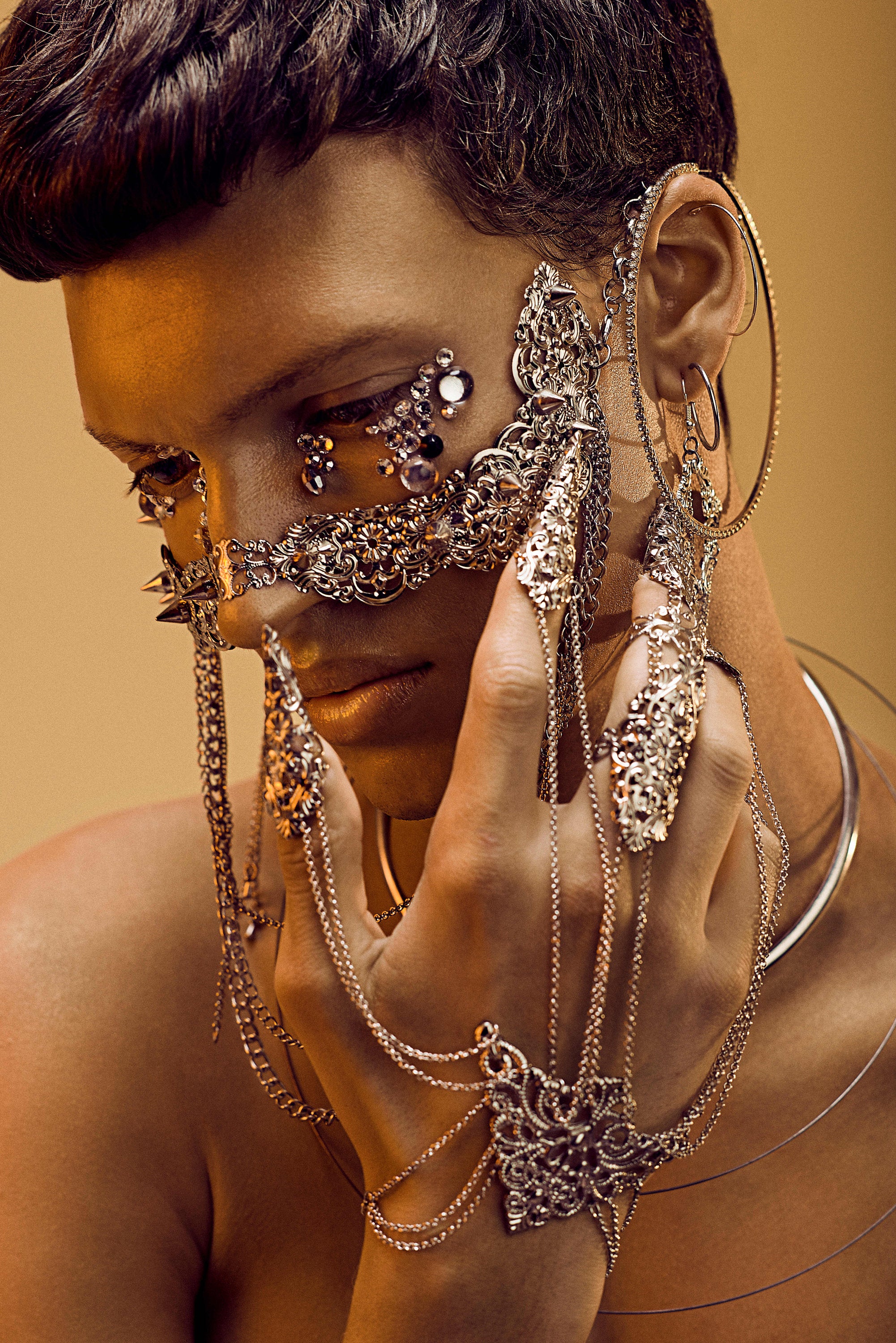 A fashion-forward male model dons a Myril Jewels studded nose mask adorned with hanging chains, presenting a luxe neo-goth look. The mask's intricate metalwork and dark-avantgarde design exemplify gothic-chic elegance, perfect for those with a penchant for unique Halloween or punk jewelry. This high-fashion piece encapsulates the whimsigoth spirit and witchcore trend, making it an audacious choice for everyday wear, rave parties, or as an eye-catching festival jewel