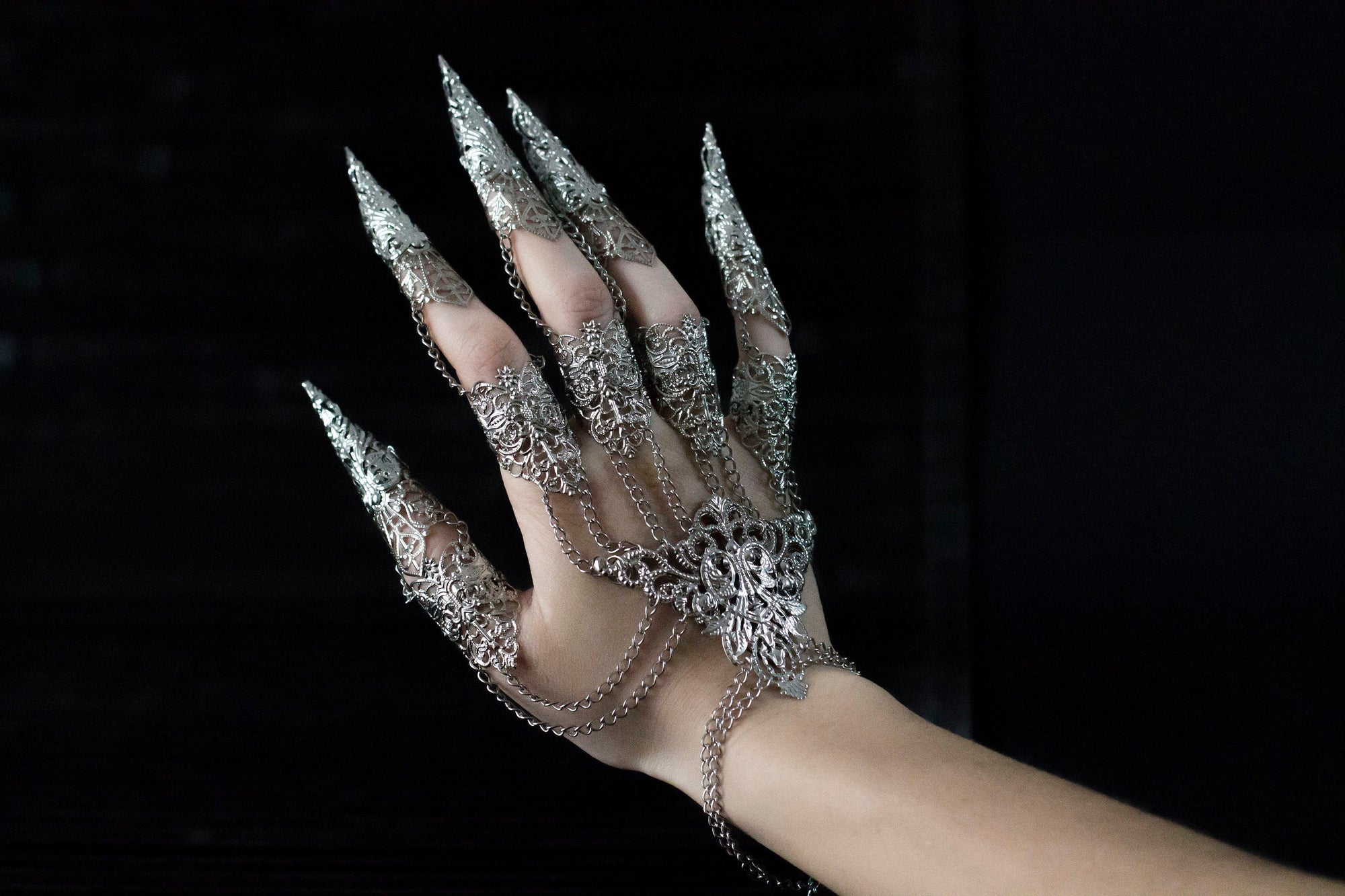 A hand adorned with an intricate silver Metal Glove Diablo, featuring detailed filigree work and sharp, elongated fingertip designs, connected by delicate chains that add a gothic elegance to the piece, set against a dark background.