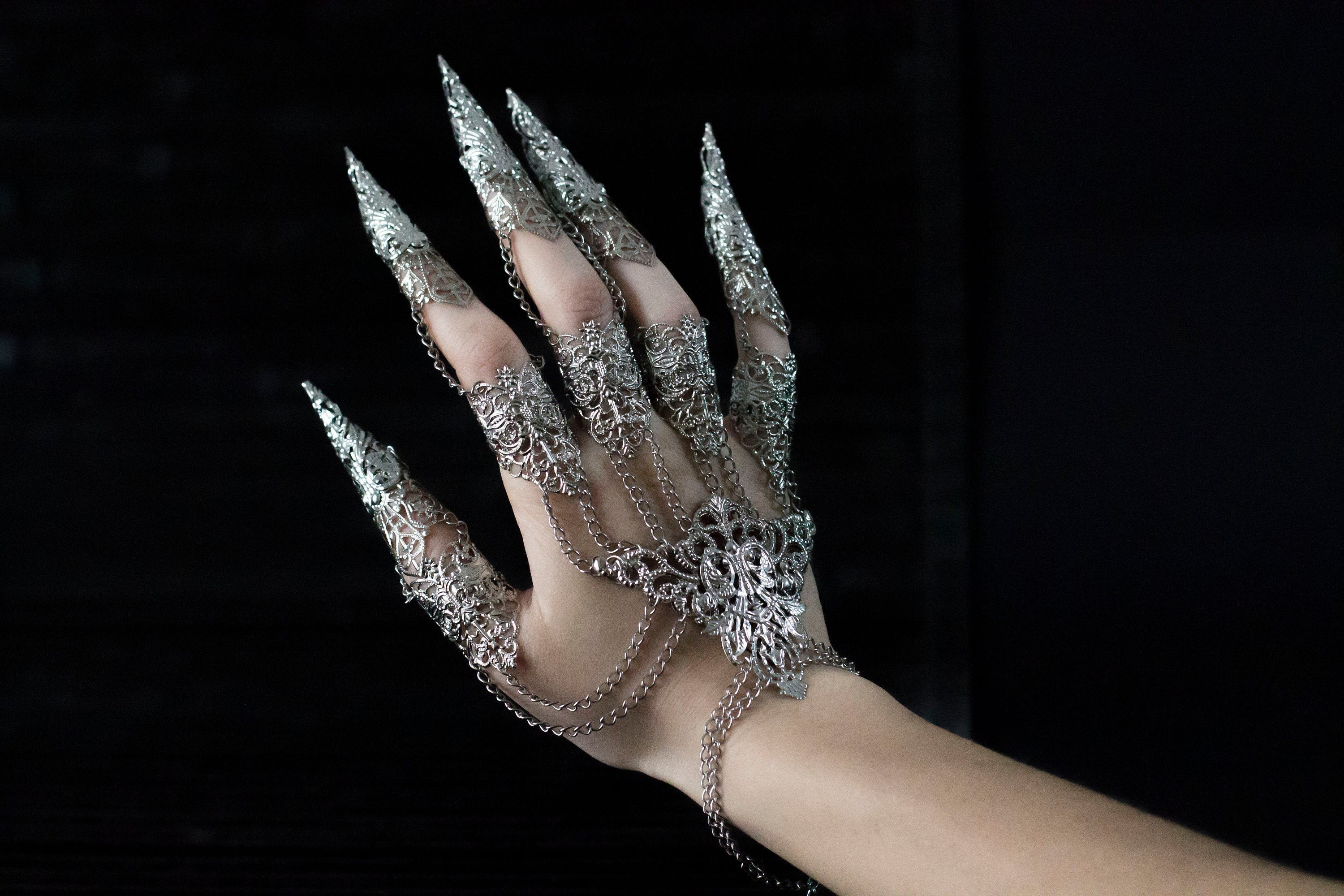 A hand adorned with an intricate silver Metal Glove Diablo, featuring detailed filigree work and sharp, elongated fingertip designs, connected by delicate chains that add a gothic elegance to the piece, set against a dark background.