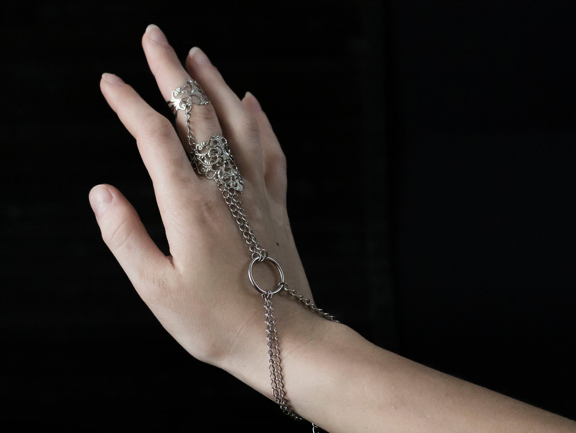 Elevate your gothic-chic style with this Myril Jewels hand chain bracelet ring, designed for the sophisticated avant-garde enthusiast. This piece features an intricate filigree design, exuding a dark, elegant allure perfect for everyday wear or a rave party statement. Crafted with the whimsigoth aesthetic in mind, the striking contrast of the shiny silver against the model's black stiletto nails encapsulates a neo-gothic vibe, making it a timeless addition to any witchcore or minimal goth jewelry collectio