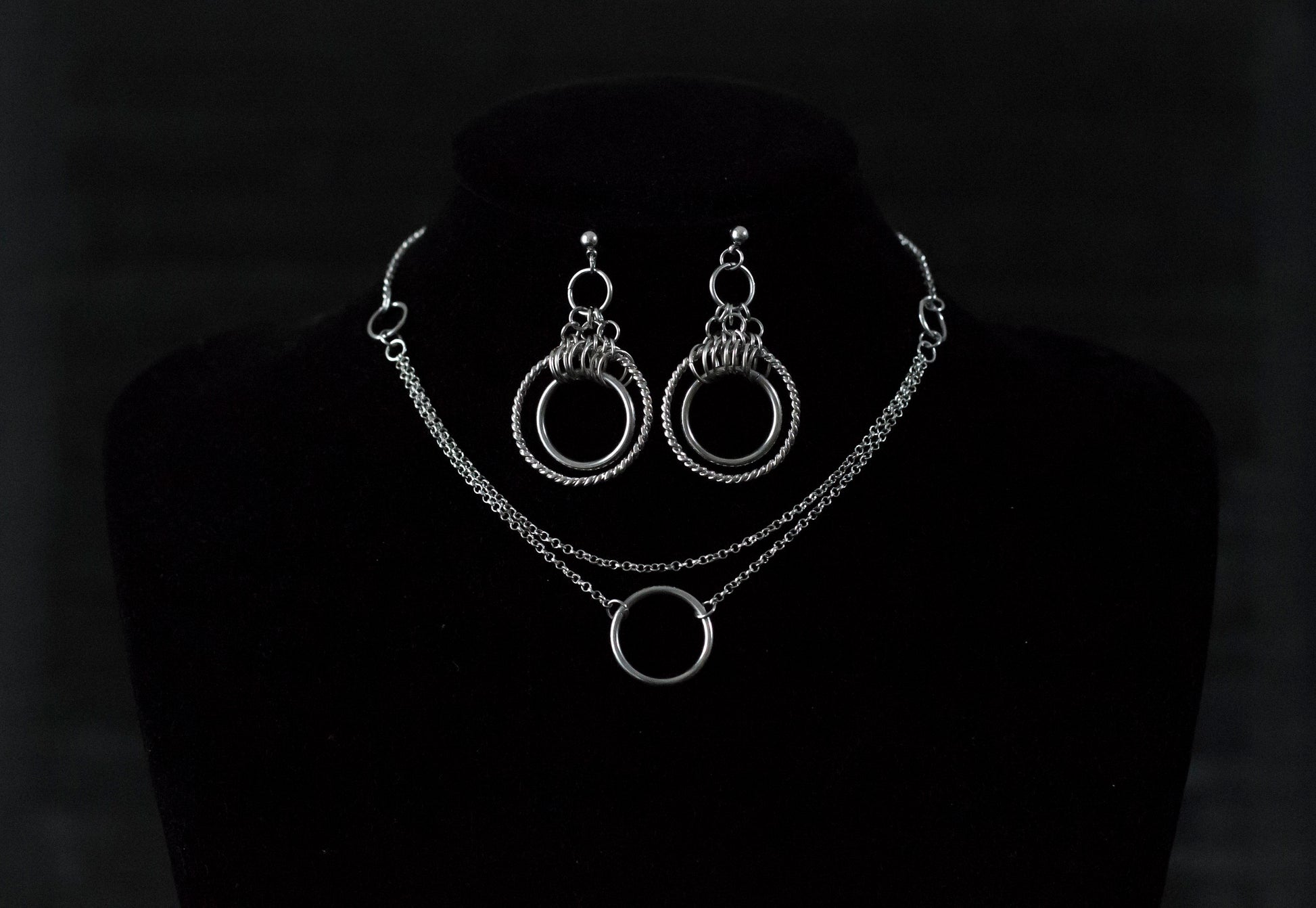 This image showcases a set of double hoop earrings and a matching O-ring chain necklace. The jewelry is displayed against a matte black mannequin bust, highlighting the silver sheen of the metalwork. The earrings feature two concentric hoops with decorative elements on the outer ring, while the necklace has a simple, elegant O-ring centerpiece connected by multiple chains of varying lengths.
