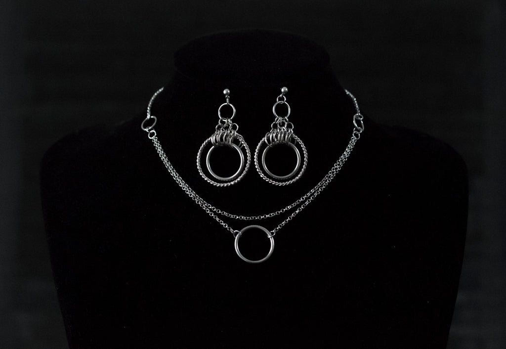 This image showcases a set of double hoop earrings and a matching O-ring chain necklace. The jewelry is displayed against a matte black mannequin bust, highlighting the silver sheen of the metalwork. The earrings feature two concentric hoops with decorative elements on the outer ring, while the necklace has a simple, elegant O-ring centerpiece connected by multiple chains of varying lengths.