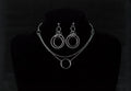 This image showcases a set of double hoop earrings and a matching O-ring chain necklace. The jewelry is displayed against a matte black mannequin bust, highlighting the silver sheen of the metalwork. The earrings feature two concentric hoops with decorative elements on the outer ring, while the necklace has a simple, elegant O-ring centerpiece connected by multiple chains of varying lengths.