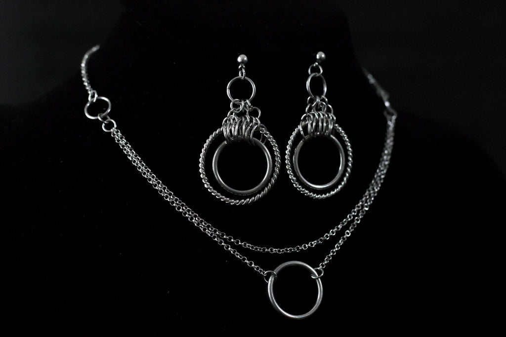 This image showcases a set of double hoop earrings and a matching O-ring chain necklace. The jewelry is displayed against a matte black mannequin bust, highlighting the silver sheen of the metalwork. The earrings feature two concentric hoops with decorative elements on the outer ring, while the necklace has a simple, elegant O-ring centerpiece connected by multiple chains of varying lengths.