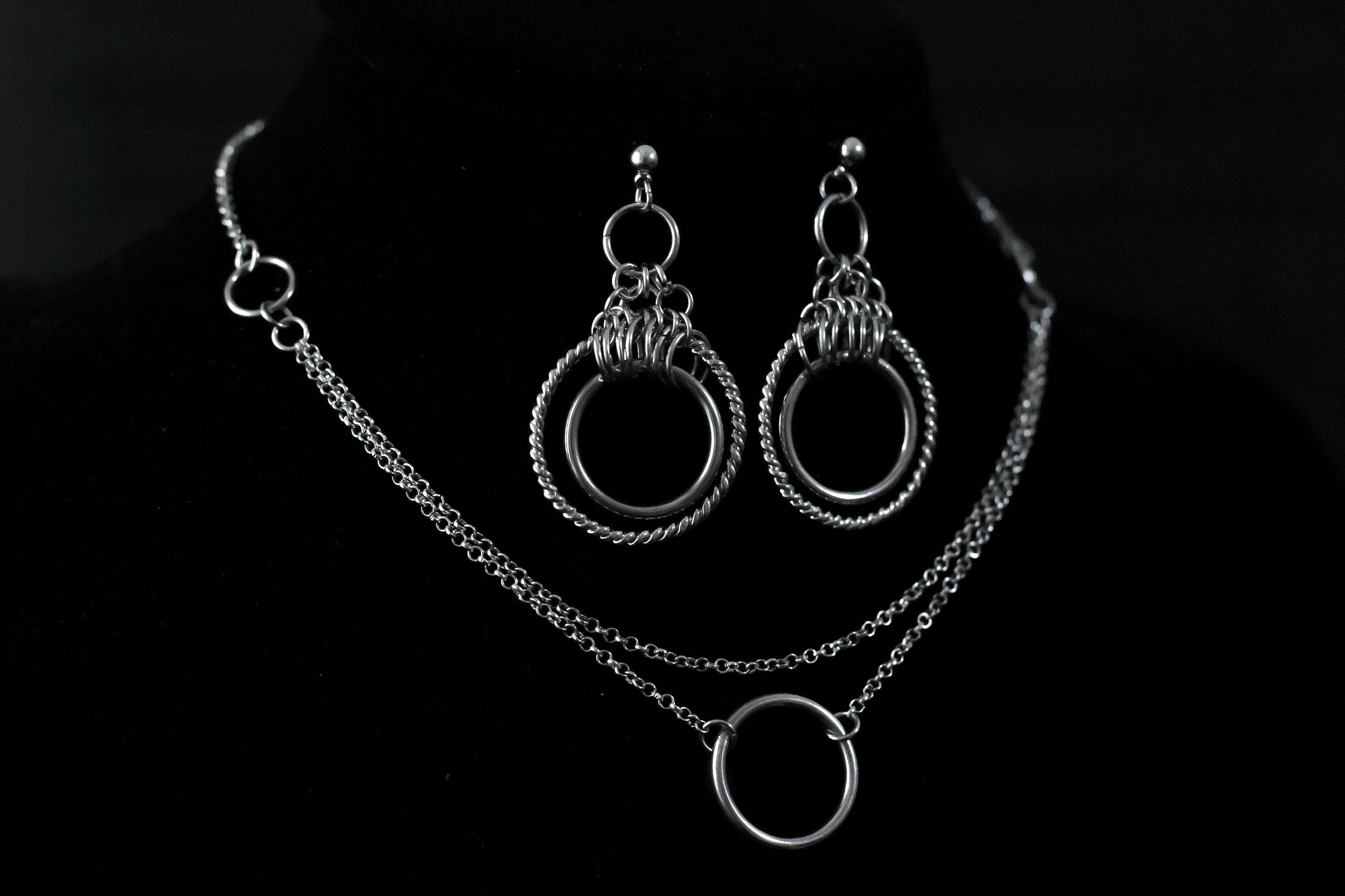 This image showcases a set of double hoop earrings and a matching O-ring chain necklace. The jewelry is displayed against a matte black mannequin bust, highlighting the silver sheen of the metalwork. The earrings feature two concentric hoops with decorative elements on the outer ring, while the necklace has a simple, elegant O-ring centerpiece connected by multiple chains of varying lengths.