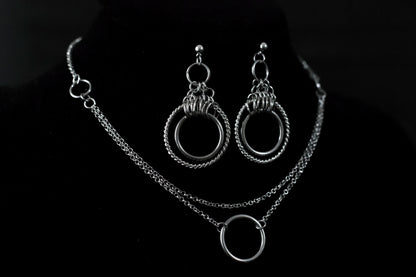 This image showcases a set of double hoop earrings and a matching O-ring chain necklace. The jewelry is displayed against a matte black mannequin bust, highlighting the silver sheen of the metalwork. The earrings feature two concentric hoops with decorative elements on the outer ring, while the necklace has a simple, elegant O-ring centerpiece connected by multiple chains of varying lengths.
