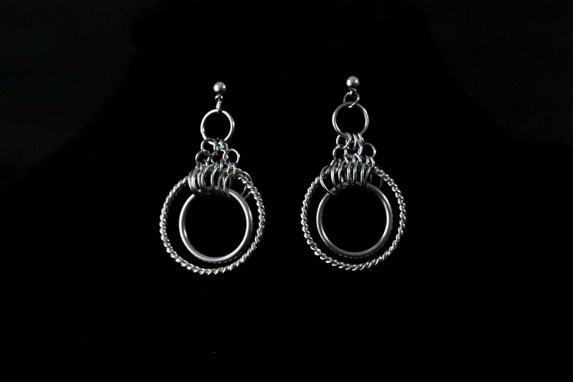 A pair of double hoop earrings, elegantly crafted in a silver tone, with a smaller hoop nestled within a larger one, both connected by a set of intricate chain links. These Myril Jewels earrings encapsulate a neo-gothic charm, ideal for those seeking a statement piece for Halloween, punk, or everyday gothic-chic ensembles. They are a standout addition to any festival outfit or as a bold jewelry gift for a goth girlfriend.
