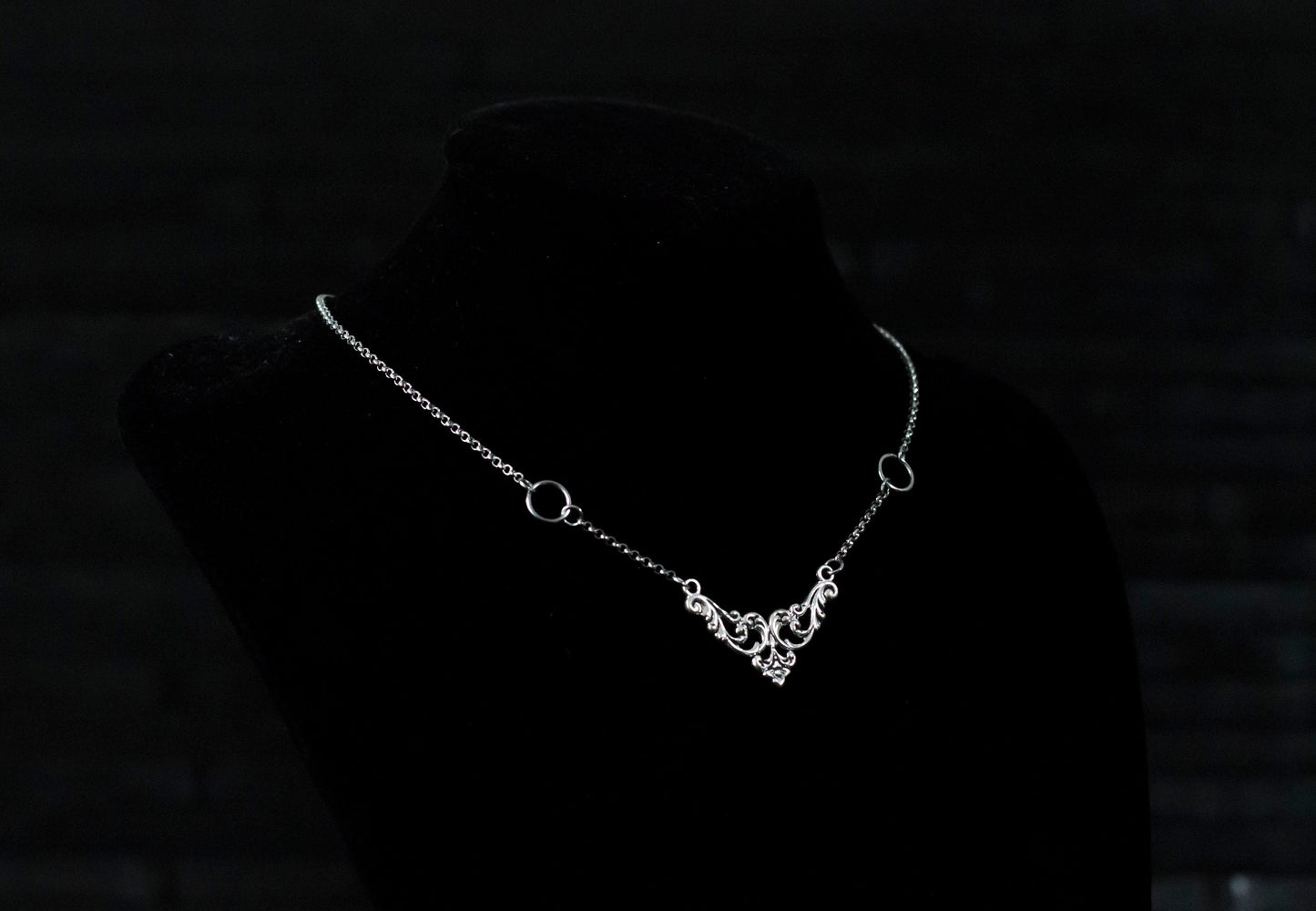 Displayed on a black velvet mannequin is a Myril Jewels elegant gothic necklace, capturing the essence of neo-goth fashion. The necklace, with a fine silver chain, features ornate filigree detailing and symmetrical O-ring accents, embodying the dark-avantgarde aesthetic. This piece is designed for gothic and alternative style aficionados, seamlessly blending with Halloween, punk, and gothic-chic ensembles, and is a sophisticated choice for whimsigoth, witchcore, or minimal goth everyday wear