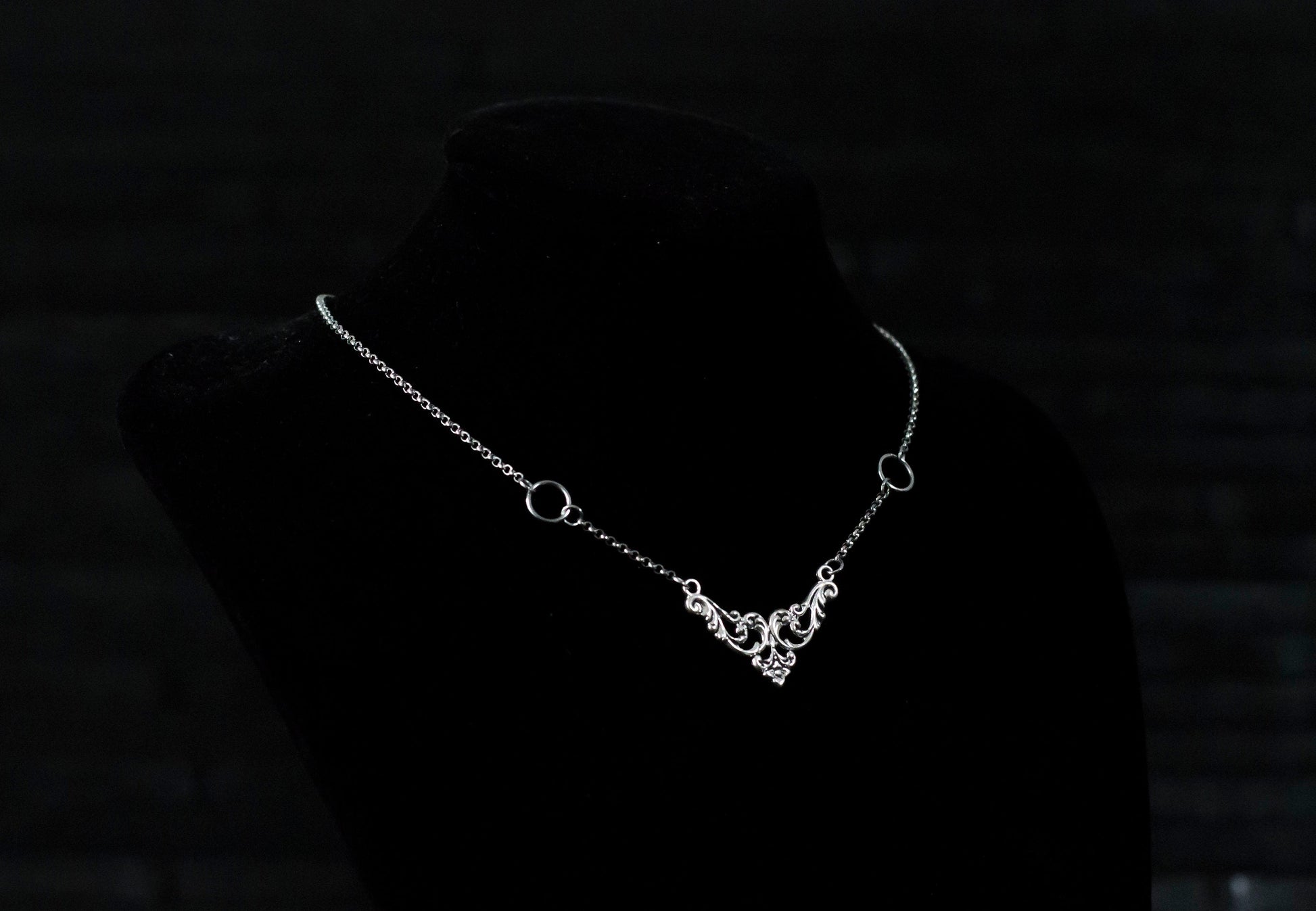 Displayed on a black velvet mannequin is a Myril Jewels elegant gothic necklace, capturing the essence of neo-goth fashion. The necklace, with a fine silver chain, features ornate filigree detailing and symmetrical O-ring accents, embodying the dark-avantgarde aesthetic. This piece is designed for gothic and alternative style aficionados, seamlessly blending with Halloween, punk, and gothic-chic ensembles, and is a sophisticated choice for whimsigoth, witchcore, or minimal goth everyday wear