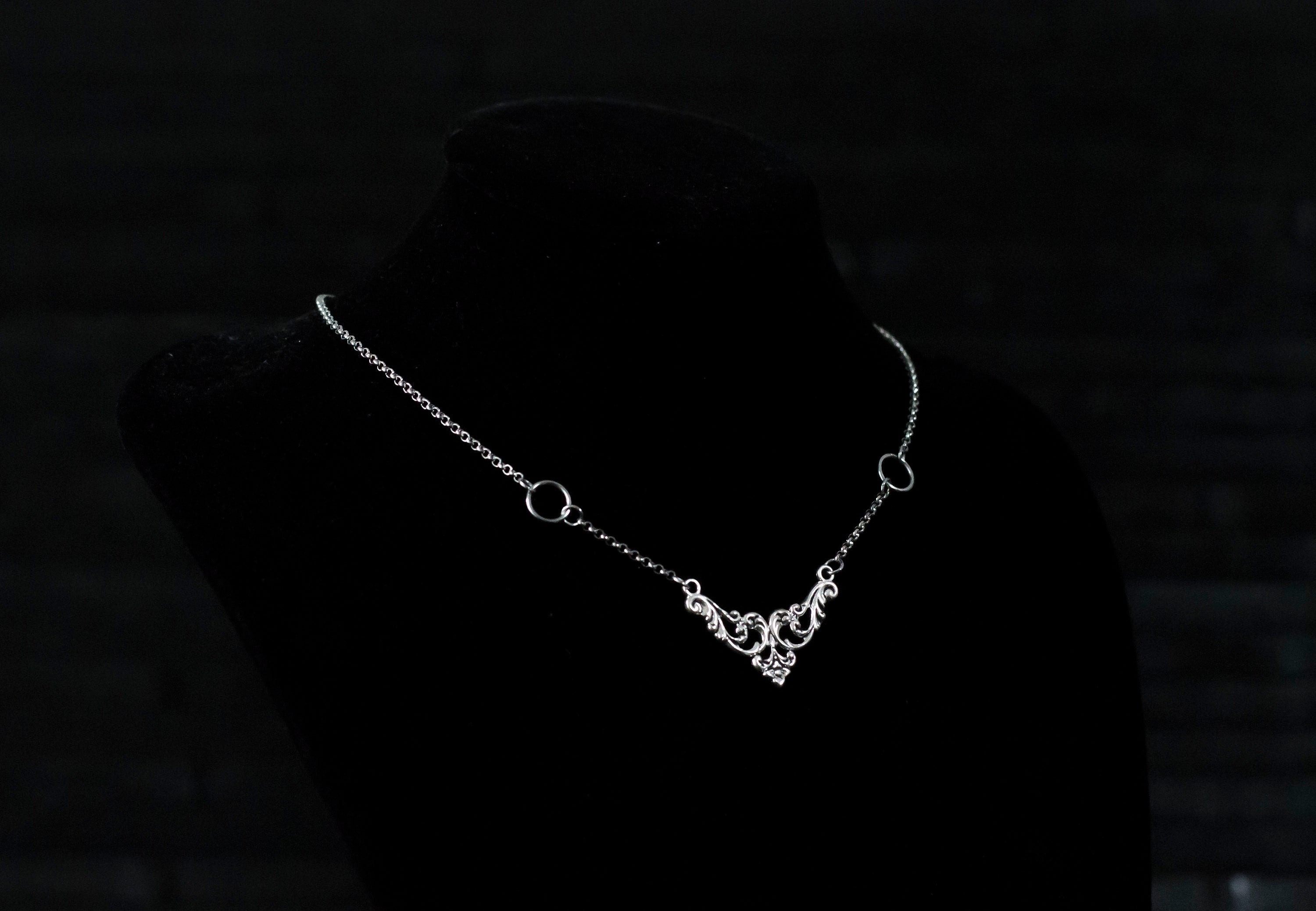 Displayed on a black velvet mannequin is a Myril Jewels elegant gothic necklace, capturing the essence of neo-goth fashion. The necklace, with a fine silver chain, features ornate filigree detailing and symmetrical O-ring accents, embodying the dark-avantgarde aesthetic. This piece is designed for gothic and alternative style aficionados, seamlessly blending with Halloween, punk, and gothic-chic ensembles, and is a sophisticated choice for whimsigoth, witchcore, or minimal goth everyday wear