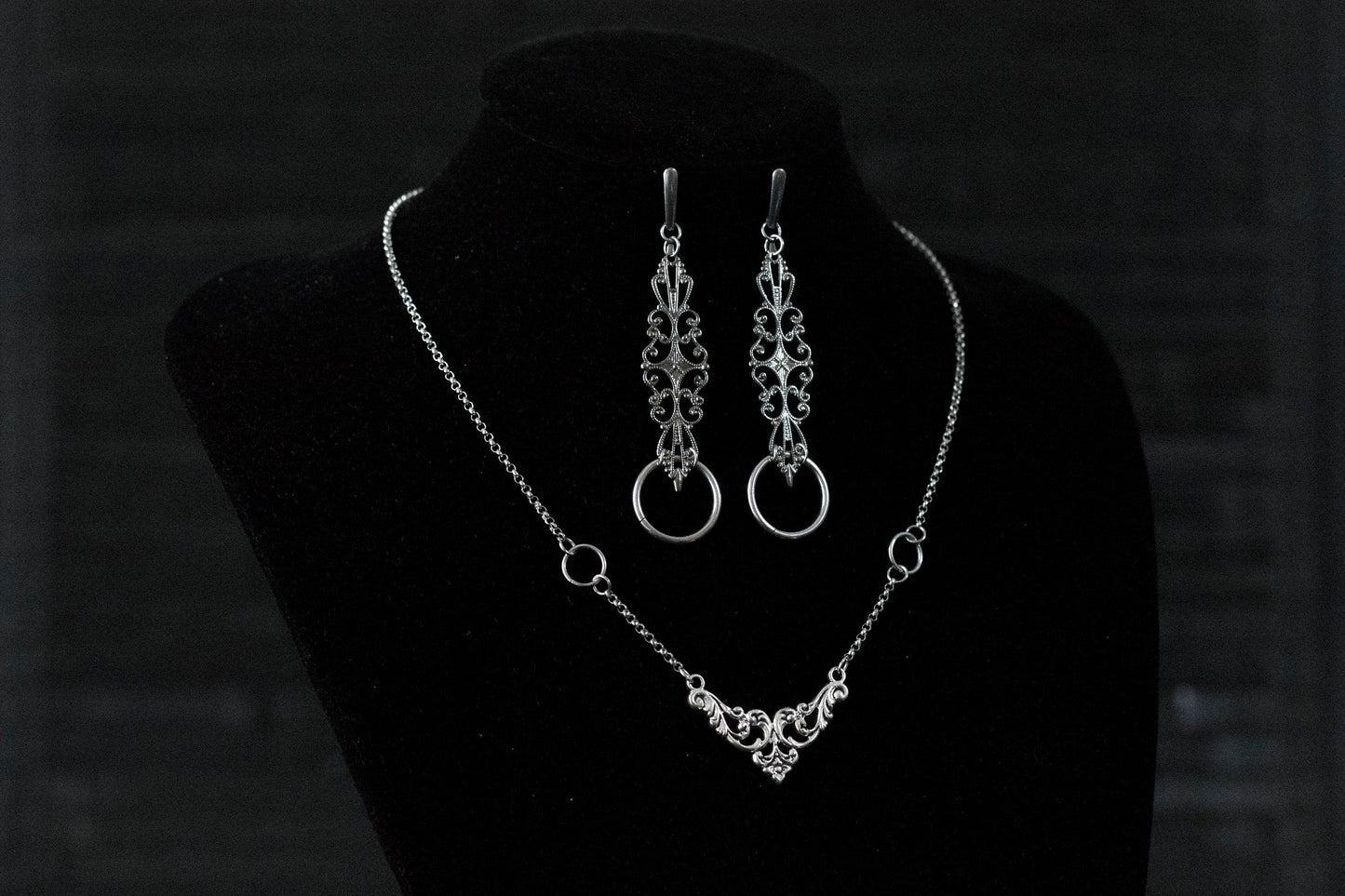 A Myril Jewels gothic necklace and matching earrings set is elegantly displayed on a black velvet mannequin. The silver necklace features a delicate filigree design, culminating in a classic gothic arch motif, paired with earrings that echo this design with an added hoop. Perfect for those who appreciate neo-goth jewelry, this set adds a touch of gothic-chic to any attire, making it ideal for Halloween, witchcore expressions, or as sophisticated everyday wear for the avant-garde individual.