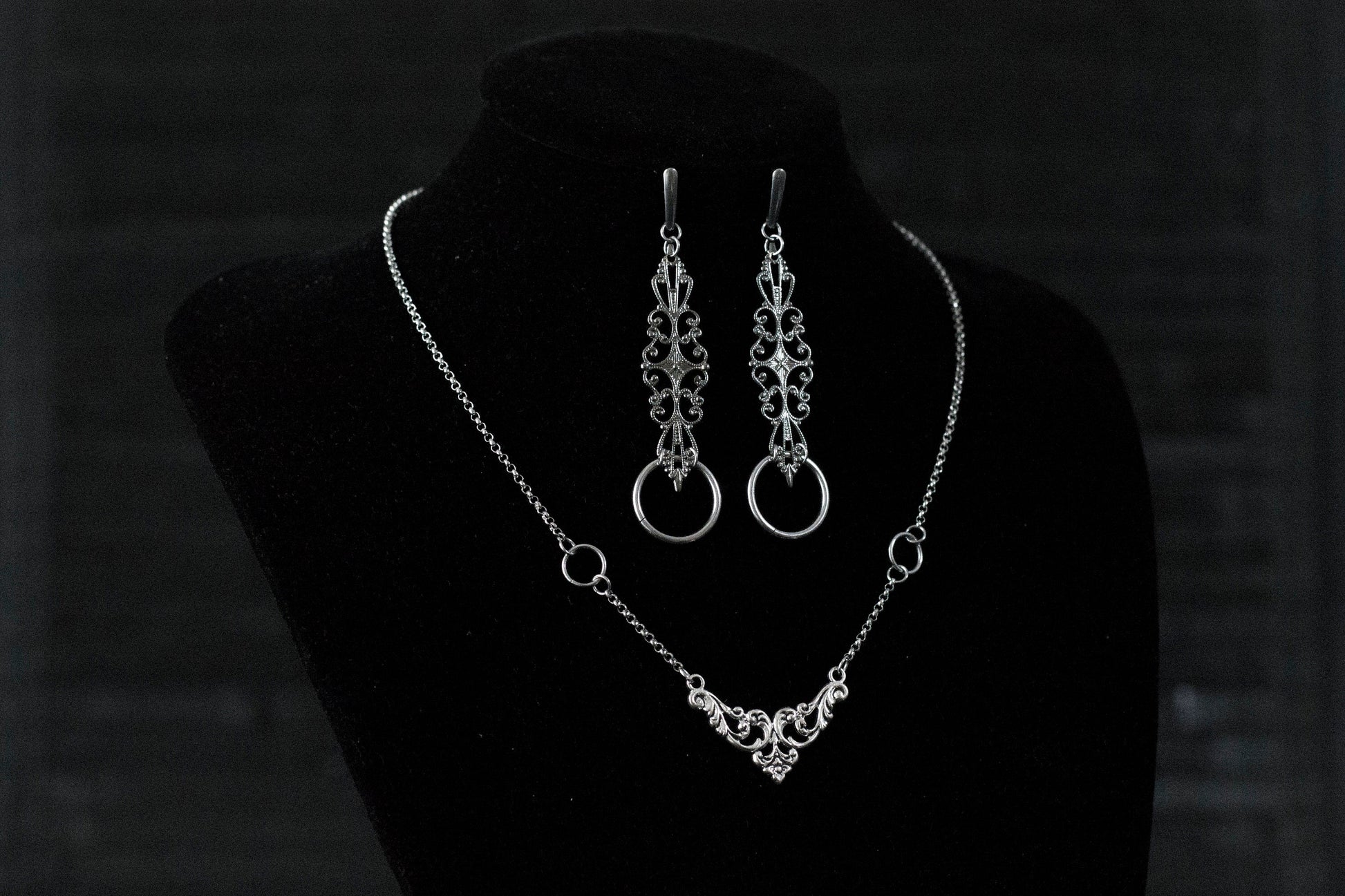 A Myril Jewels gothic necklace and matching earrings set is elegantly displayed on a black velvet mannequin. The silver necklace features a delicate filigree design, culminating in a classic gothic arch motif, paired with earrings that echo this design with an added hoop. Perfect for those who appreciate neo-goth jewelry, this set adds a touch of gothic-chic to any attire, making it ideal for Halloween, witchcore expressions, or as sophisticated everyday wear for the avant-garde individual.