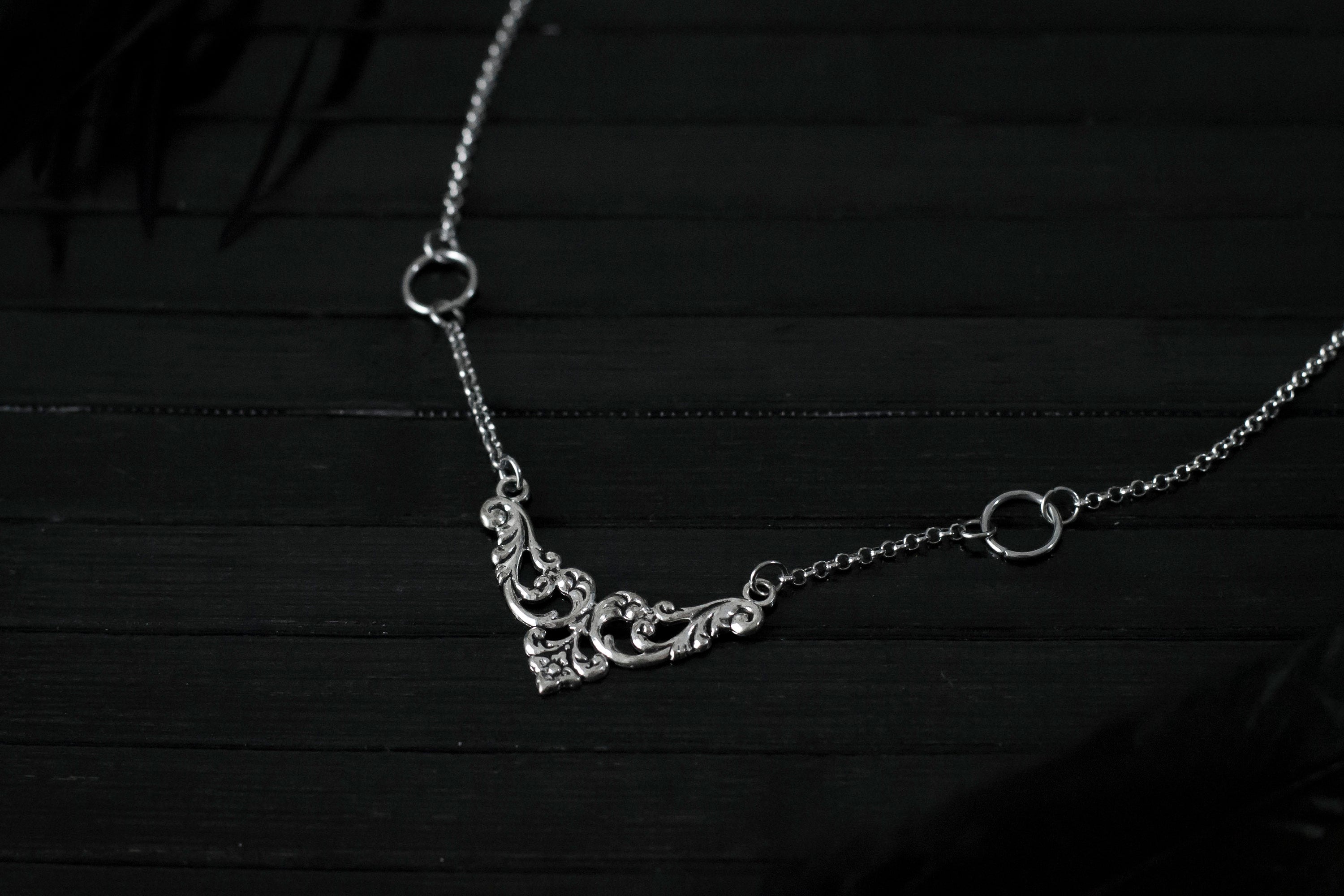 Displayed on a black layer is a Myril Jewels elegant gothic necklace, capturing the essence of neo-goth fashion. The necklace, with a fine silver chain, features ornate filigree detailing and symmetrical O-ring accents, embodying the dark-avantgarde aesthetic. This piece is designed for gothic and alternative style aficionados, seamlessly blending with Halloween, punk, and gothic-chic ensembles, and is a sophisticated choice for whimsigoth, witchcore, or minimal goth everyday wear