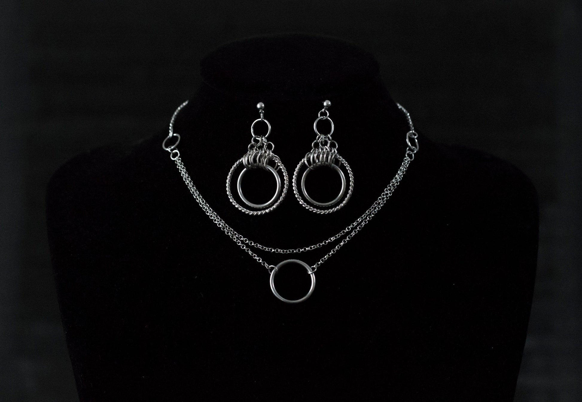 This image showcases a set of double hoop earrings and a matching O-ring chain necklace. The jewelry is displayed against a matte black mannequin bust, highlighting the silver sheen of the metalwork. The earrings feature two concentric hoops with decorative elements on the outer ring, while the necklace has a simple, elegant O-ring centerpiece connected by multiple chains of varying lengths.