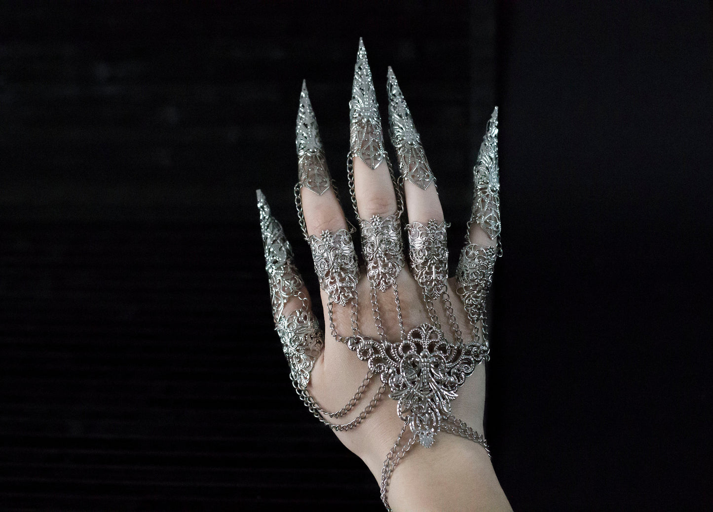 A hand adorned with an intricate silver Metal Glove Diablo, featuring detailed filigree work and sharp, elongated fingertip designs, connected by delicate chains that add a gothic elegance to the piece, set against a dark background.