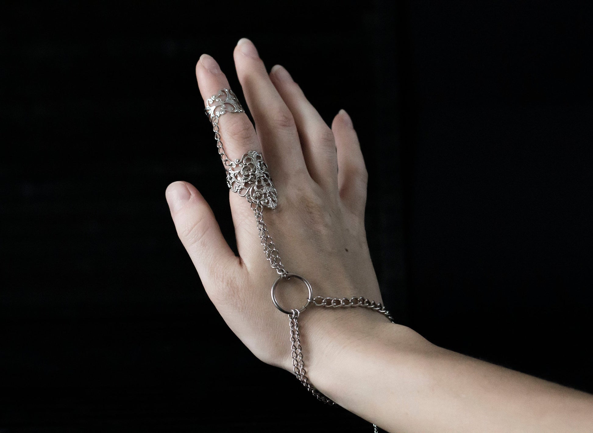 Experience the edgy allure of Myril Jewels with this handcrafted punk hand chain bracelet ring, a perfect complement to gothic and alternative styles. This piece features a lustrous, interwoven chain that drapes elegantly from a central ring to a detailed filigree band, wrapping around the middle finger for a secure fit. Its intricate design captures the essence of dark-avantgarde aesthetics, making it a must-have for those who resonate with neo-gothic charm and whimsigoth elegance.