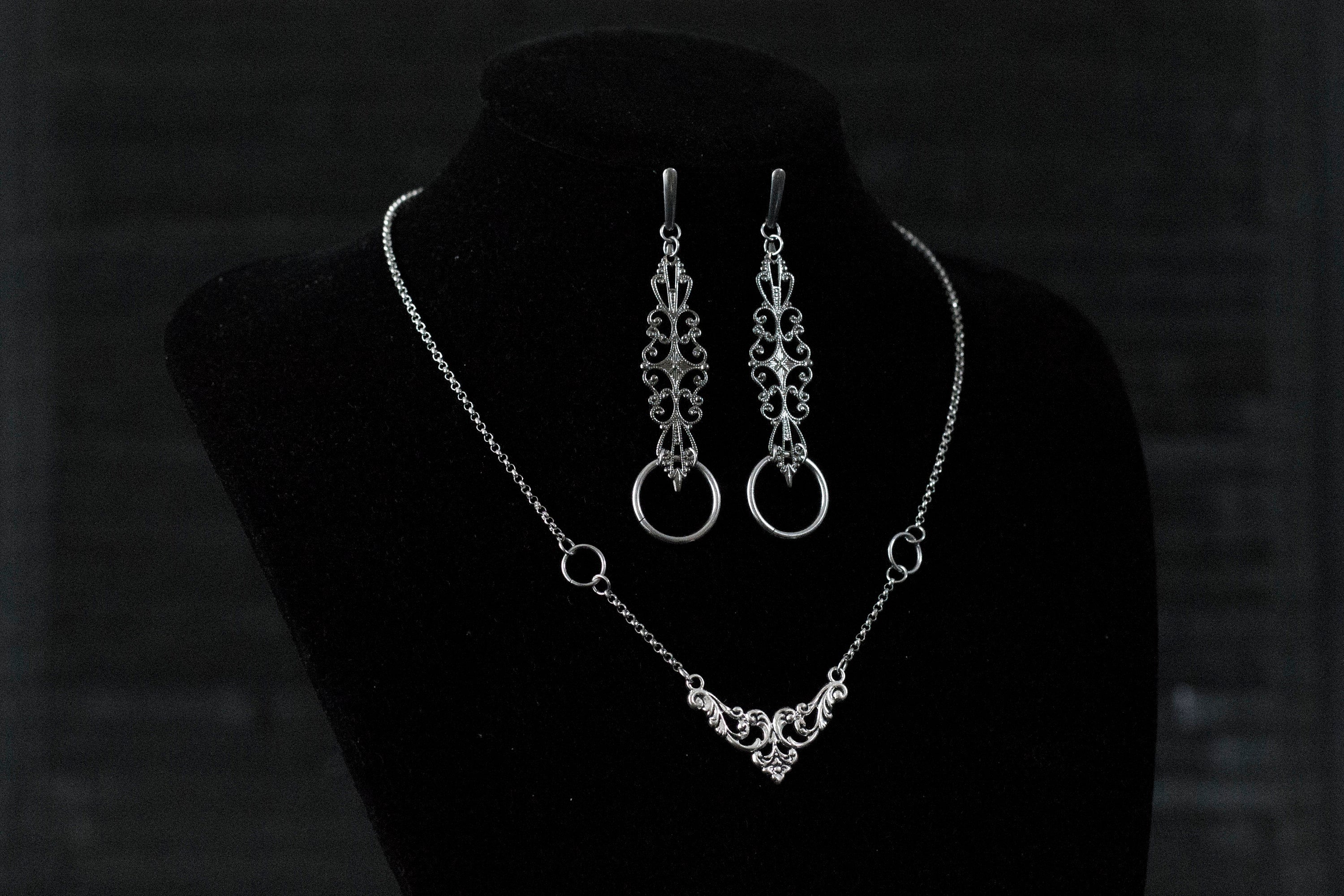 A Myril Jewels gothic necklace and matching earrings set is elegantly displayed on a black velvet mannequin. The silver necklace features a delicate filigree design, culminating in a classic gothic arch motif, paired with earrings that echo this design with an added hoop. Perfect for those who appreciate neo-goth jewelry, this set adds a touch of gothic-chic to any attire, making it ideal for Halloween, witchcore expressions, or as sophisticated everyday wear for the avant-garde individual.