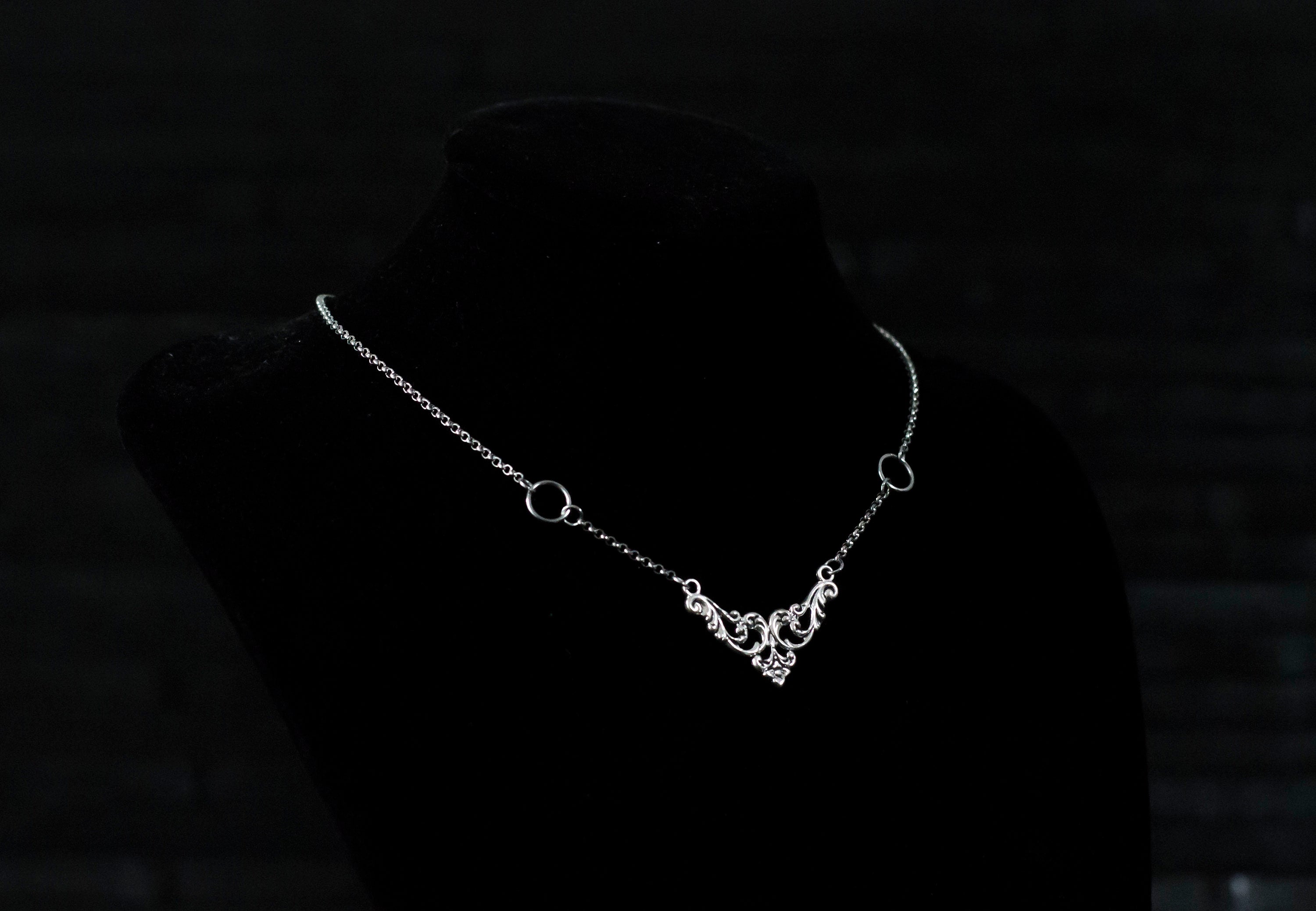 Displayed on a black velvet mannequin is a Myril Jewels elegant gothic necklace, capturing the essence of neo-goth fashion. The necklace, with a fine silver chain, features ornate filigree detailing and symmetrical O-ring accents, embodying the dark-avantgarde aesthetic. This piece is designed for gothic and alternative style aficionados, seamlessly blending with Halloween, punk, and gothic-chic ensembles, and is a sophisticated choice for whimsigoth, witchcore, or minimal goth everyday wear