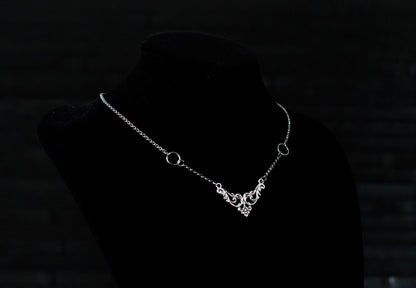 Displayed on a black velvet mannequin is a Myril Jewels elegant gothic necklace, capturing the essence of neo-goth fashion. The necklace, with a fine silver chain, features ornate filigree detailing and symmetrical O-ring accents, embodying the dark-avantgarde aesthetic. This piece is designed for gothic and alternative style aficionados, seamlessly blending with Halloween, punk, and gothic-chic ensembles, and is a sophisticated choice for whimsigoth, witchcore, or minimal goth everyday wear