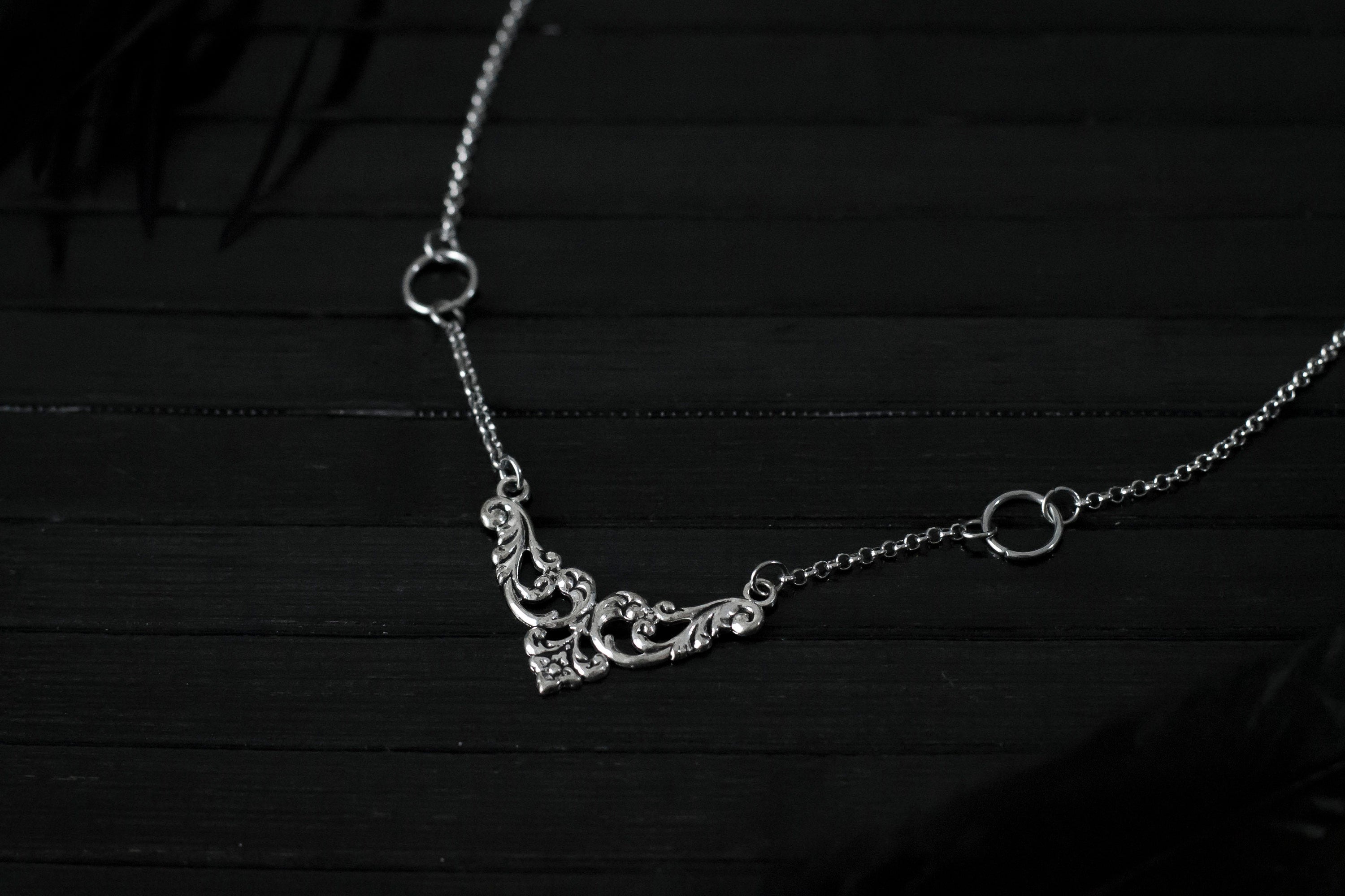 Displayed on a black layer is a Myril Jewels elegant gothic necklace, capturing the essence of neo-goth fashion. The necklace, with a fine silver chain, features ornate filigree detailing and symmetrical O-ring accents, embodying the dark-avantgarde aesthetic. This piece is designed for gothic and alternative style aficionados, seamlessly blending with Halloween, punk, and gothic-chic ensembles, and is a sophisticated choice for whimsigoth, witchcore, or minimal goth everyday wear