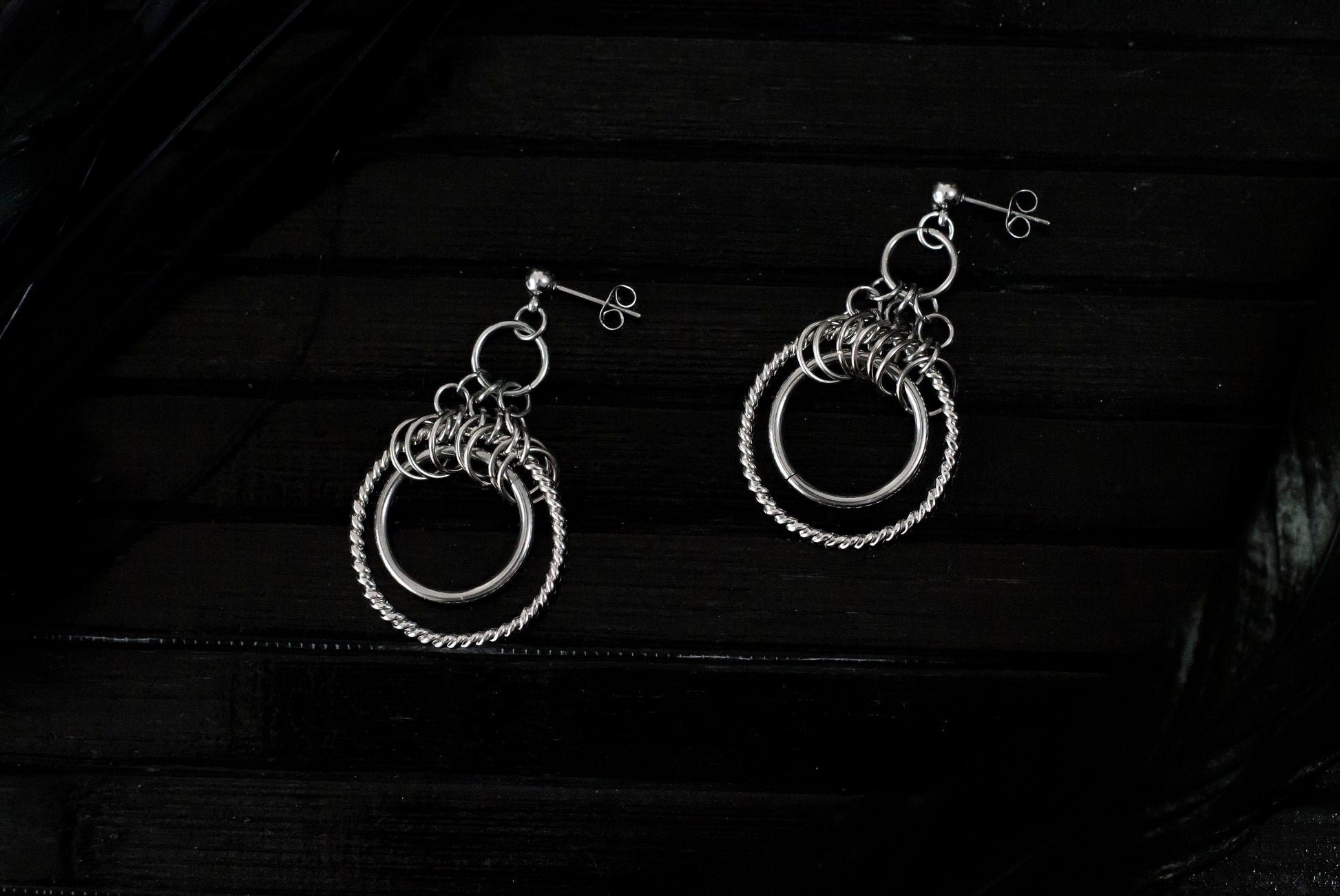 The image displays a pair of double hoop earrings with a sophisticated design. The earrings feature two concentric circles; the inner hoop is smooth and polished, while the outer hoop has a twisted rope-like texture, enhancing its visual appeal. Above the hoops, there is a cluster of smaller rings adding complexity to the design. These earrings, likely from Myril Jewels, reflect a gothic-chic style that merges elegance with an edgy touch, making them suitable for various occasions