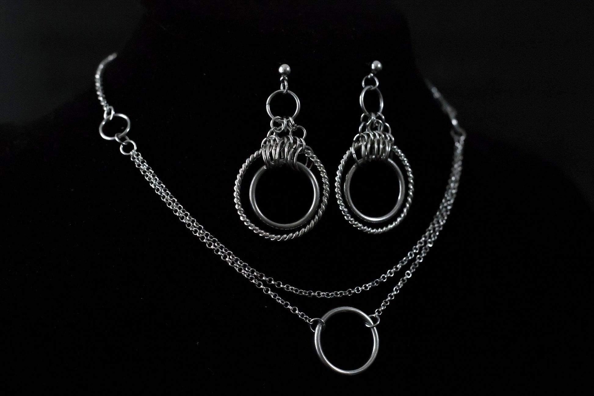 The image presents a striking set of double hoop earrings and a matching o-ring chain necklace displayed on a dark mannequin. The jewelry is characterized by its gleaming silver finish, accentuating a bold gothic design that captures the avant-garde essence of Myril Jewels. This set exemplifies the brand's dedication to crafting accessories for those who embrace a dark, gothic aesthetic, and it resonates with the themes of punk jewelry and neo-gothic style.