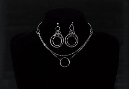 The image presents a striking set of double hoop earrings and a matching o-ring chain necklace displayed on a dark mannequin. The jewelry is characterized by its gleaming silver finish, accentuating a bold gothic design that captures the avant-garde essence of Myril Jewels. This set exemplifies the brand's dedication to crafting accessories for those who embrace a dark, gothic aesthetic, and it resonates with the themes of punk jewelry and neo-gothic style.