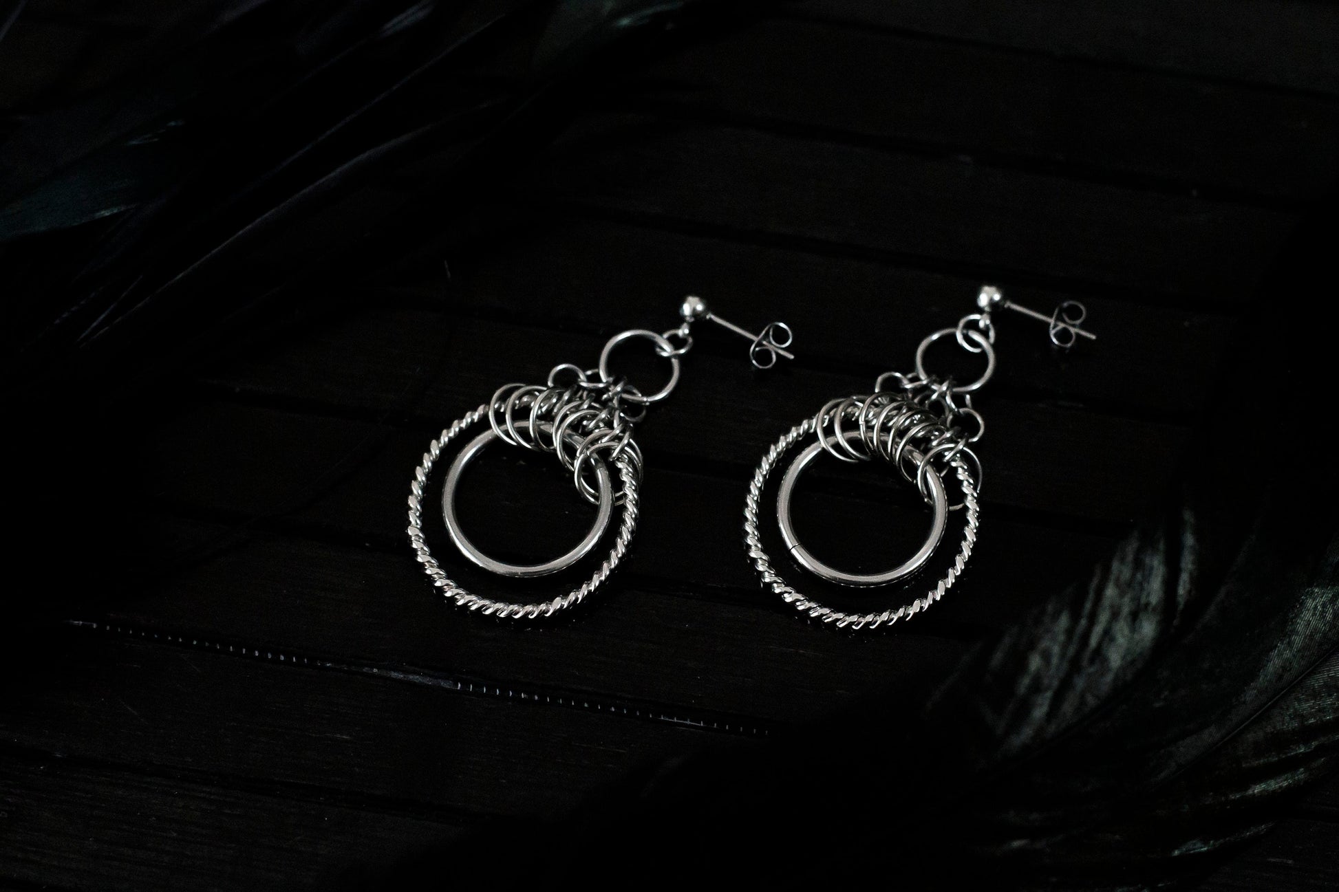 The image displays a pair of double hoop earrings with a sophisticated design. The earrings feature two concentric circles; the inner hoop is smooth and polished, while the outer hoop has a twisted rope-like texture, enhancing its visual appeal. Above the hoops, there is a cluster of smaller rings adding complexity to the design. These earrings, likely from Myril Jewels, reflect a gothic-chic style that merges elegance with an edgy touch, making them suitable for various occasions