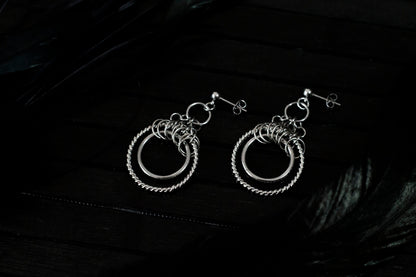 The image displays a pair of double hoop earrings with a sophisticated design. The earrings feature two concentric circles; the inner hoop is smooth and polished, while the outer hoop has a twisted rope-like texture, enhancing its visual appeal. Above the hoops, there is a cluster of smaller rings adding complexity to the design. These earrings, likely from Myril Jewels, reflect a gothic-chic style that merges elegance with an edgy touch, making them suitable for various occasions