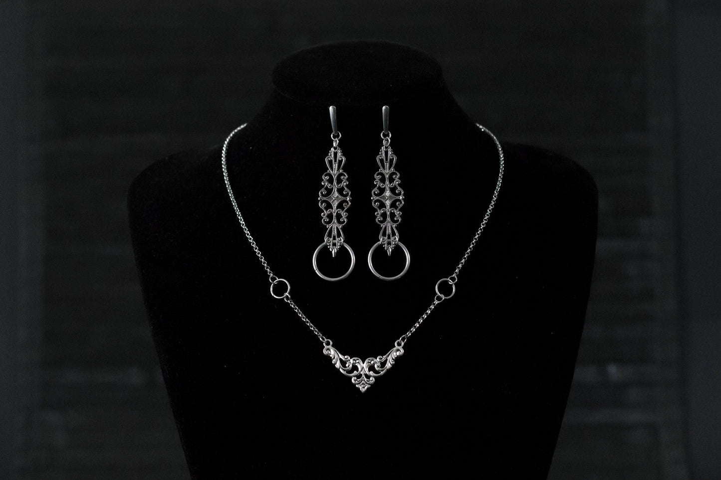 Displayed against a black backdrop is Myril Jewels' gothic necklace and earring set, exuding delicate elegance. The silver necklace features a central ornate design, flanked by symmetrical rings, reflecting neo-goth craftsmanship. Matching earrings with similar filigree patterns and rings complete this dark-avantgarde ensemble. Ideal for Halloween, this set aligns with the gothic-chic, whimsigoth, and witchcore trends, suited for both everyday wear and as a statement piece for rave parties or festivals.