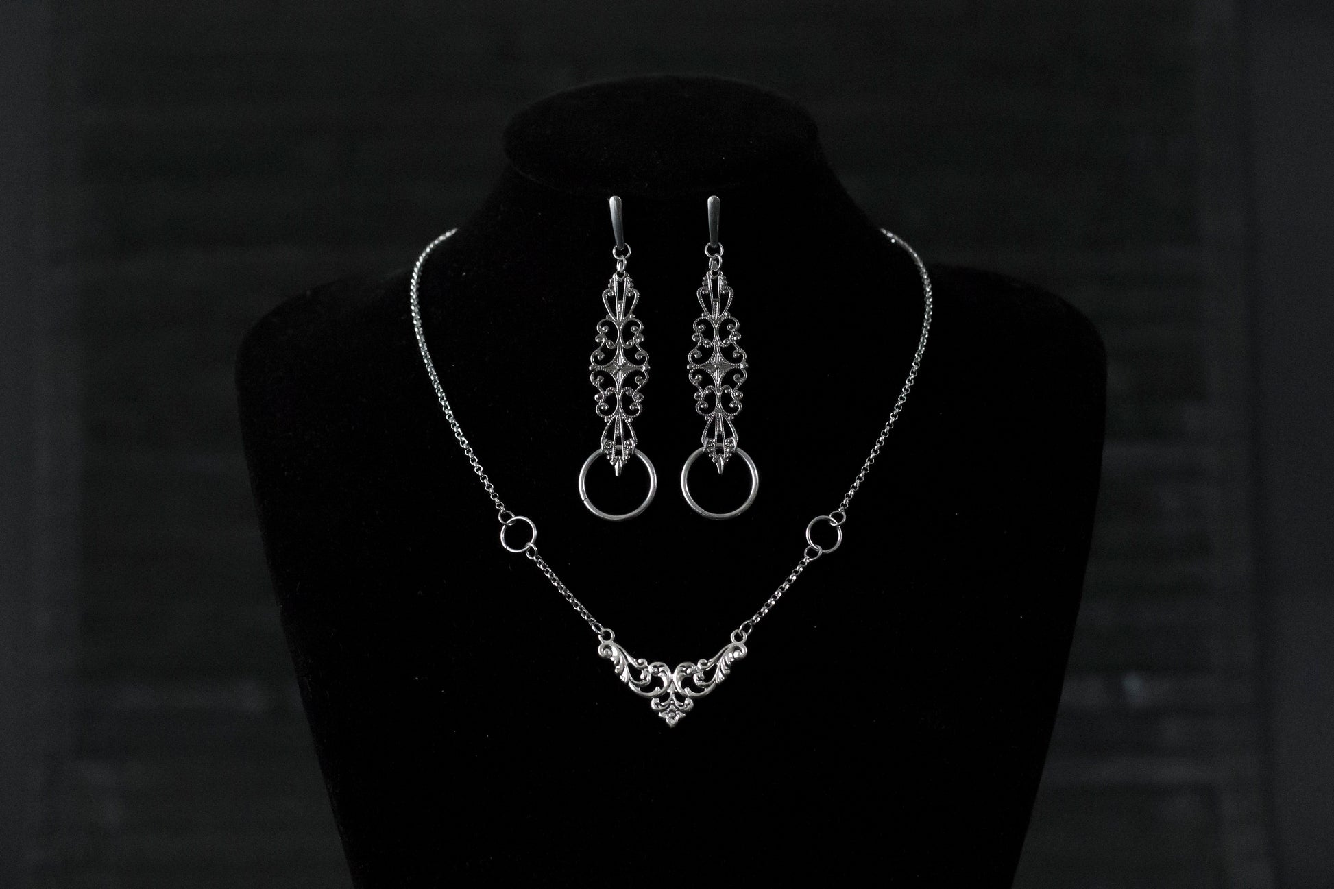Displayed against a black backdrop is Myril Jewels' gothic necklace and earring set, exuding delicate elegance. The silver necklace features a central ornate design, flanked by symmetrical rings, reflecting neo-goth craftsmanship. Matching earrings with similar filigree patterns and rings complete this dark-avantgarde ensemble. Ideal for Halloween, this set aligns with the gothic-chic, whimsigoth, and witchcore trends, suited for both everyday wear and as a statement piece for rave parties or festivals.