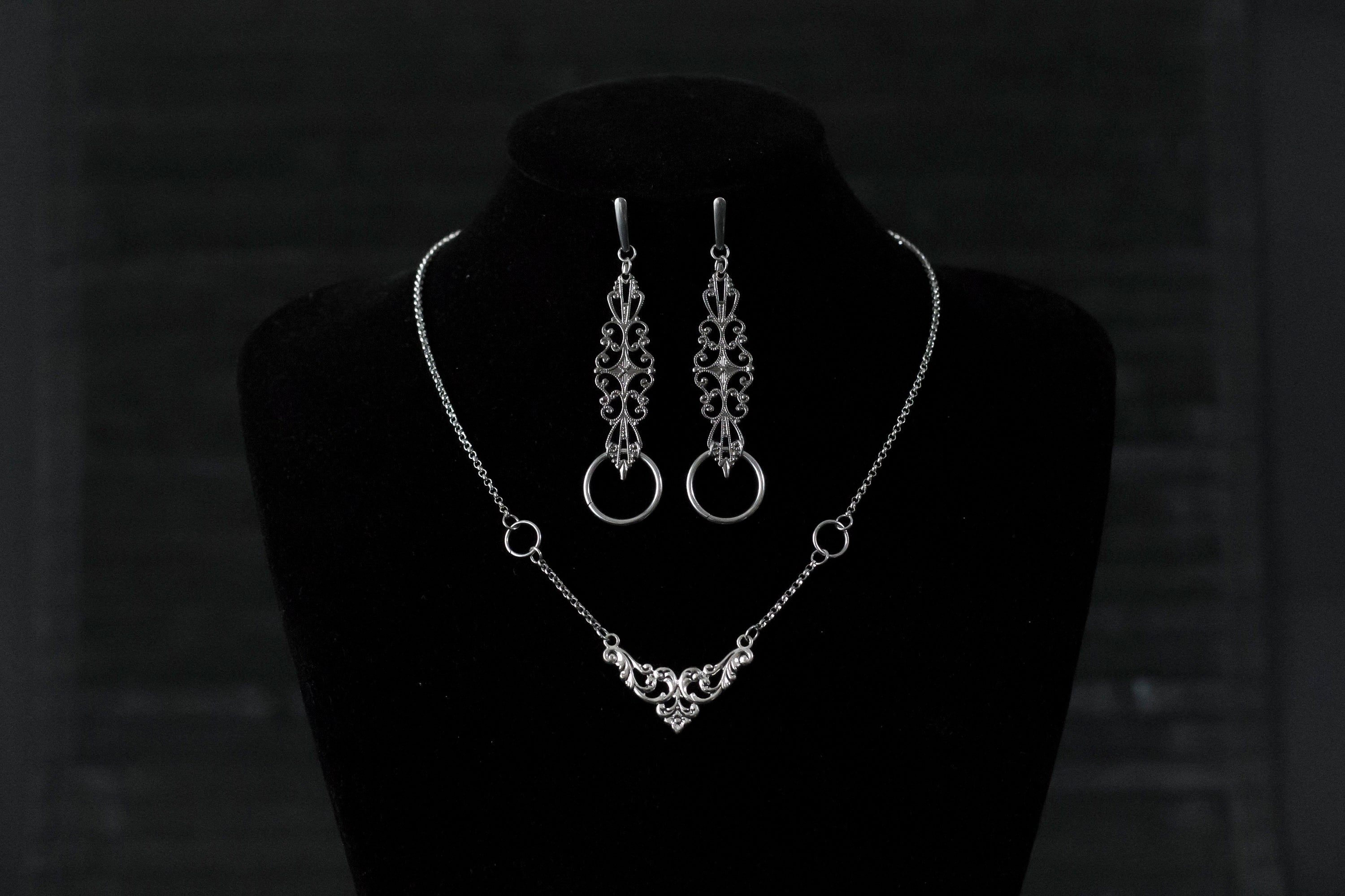 Displayed against a black backdrop is Myril Jewels' gothic necklace and earring set, exuding delicate elegance. The silver necklace features a central ornate design, flanked by symmetrical rings, reflecting neo-goth craftsmanship. Matching earrings with similar filigree patterns and rings complete this dark-avantgarde ensemble. Ideal for Halloween, this set aligns with the gothic-chic, whimsigoth, and witchcore trends, suited for both everyday wear and as a statement piece for rave parties or festivals.