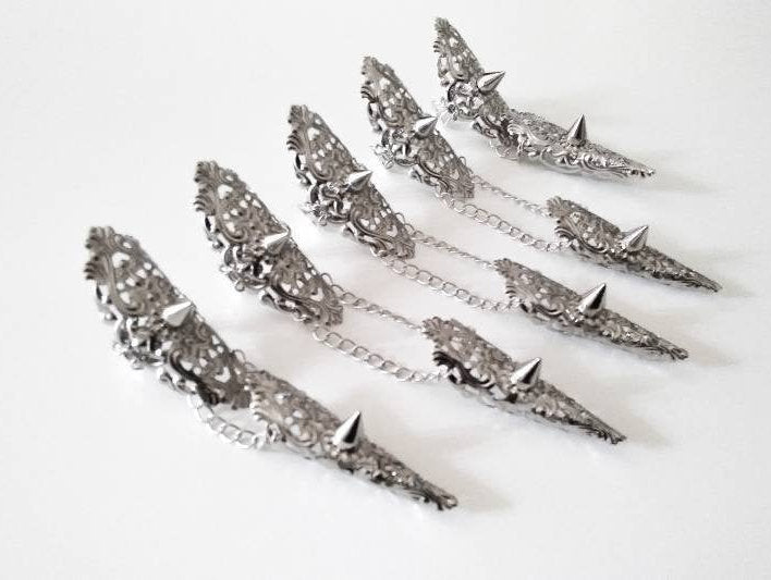 Displayed are Myril Jewels' handcrafted studded nail claw rings, laid out in a still life arrangement. These silver rings, each connected by a delicate chain, are adorned with intricate filigree and pointed studs, reflecting the brand's dedication to dark-avantgarde and neo-gothic design. They are ideal for anyone seeking a bold, punk-inspired look, and suitable for enhancing Halloween costumes or adding an edgy flair to everyday gothic-chic and witchcore fashion
