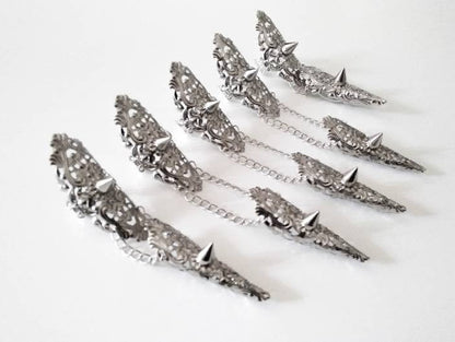 Displayed are Myril Jewels' handcrafted studded nail claw rings, laid out in a still life arrangement. These silver rings, each connected by a delicate chain, are adorned with intricate filigree and pointed studs, reflecting the brand's dedication to dark-avantgarde and neo-gothic design. They are ideal for anyone seeking a bold, punk-inspired look, and suitable for enhancing Halloween costumes or adding an edgy flair to everyday gothic-chic and witchcore fashion