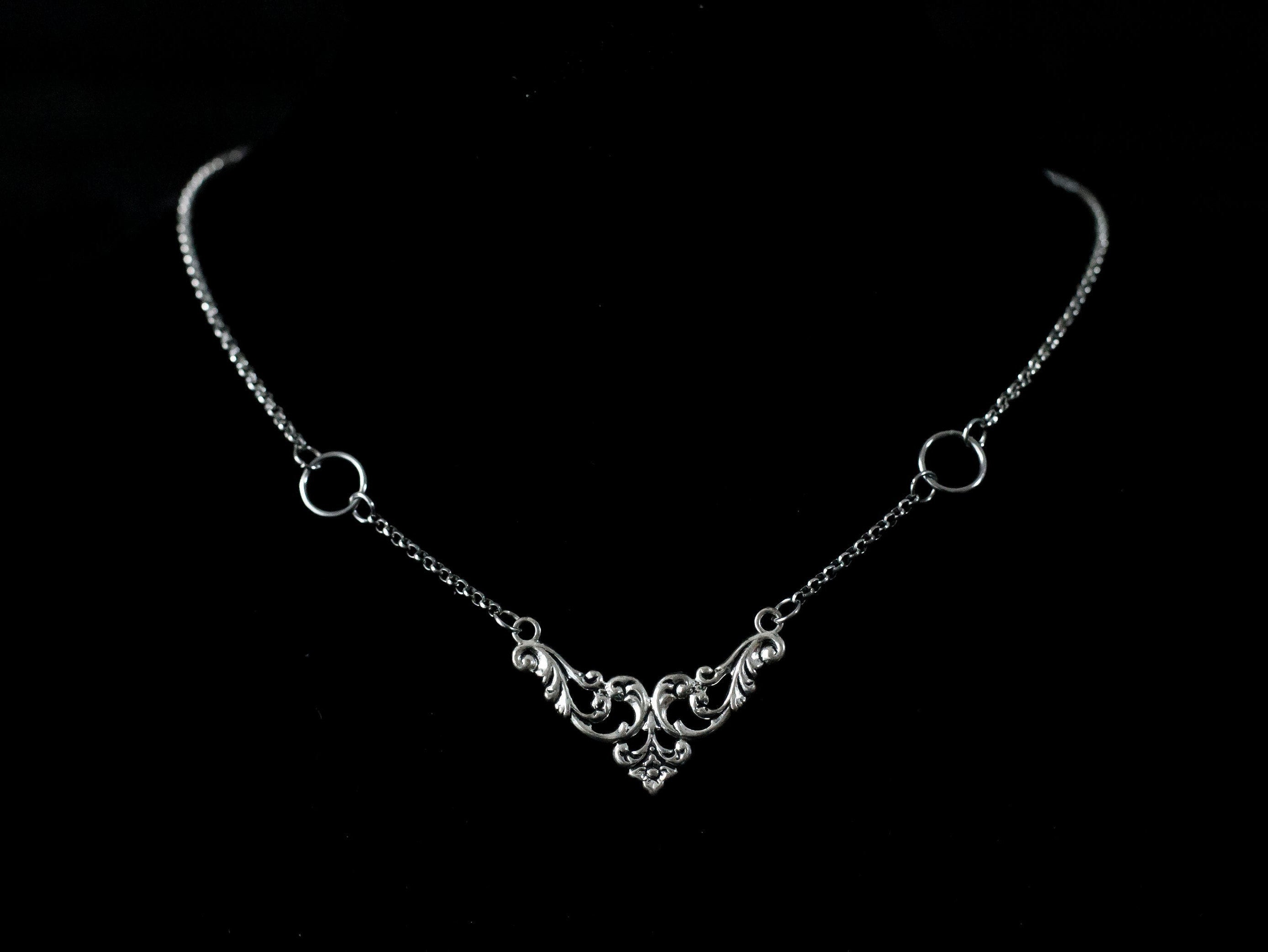 Displayed on a black velvet mannequin is a Myril Jewels elegant gothic necklace, capturing the essence of neo-goth fashion. The necklace, with a fine silver chain, features ornate filigree detailing and symmetrical O-ring accents, embodying the dark-avantgarde aesthetic. This piece is designed for gothic and alternative style aficionados, seamlessly blending with Halloween, punk, and gothic-chic ensembles, and is a sophisticated choice for whimsigoth, witchcore, or minimal goth everyday wear