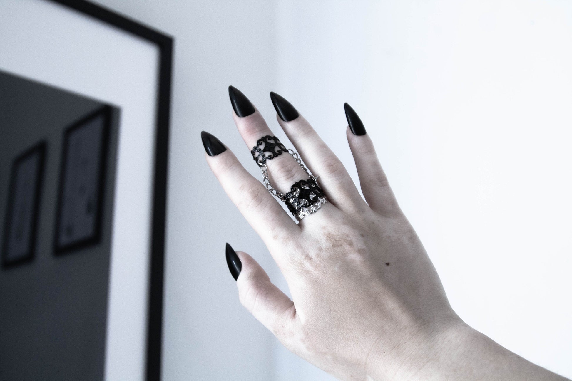  A hand with long black stiletto nails wears an intricately designed Myril Jewels double ring, connected by a delicate chain, embodies neo-gothic finesse. Perfect for witchcore fashion, this piece is an edgy accessory for gothic-chic style, making it an ideal gift for the bold goth girlfriend or as a standout festival jewel.