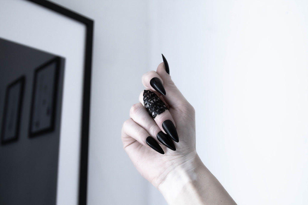 A captivating Myril Jewels goth midi ring adorns a hand with striking black polished nails, embodying the essence of neo-gothic charm. The intricate design, reminiscent of delicate cobwebs, pairs exquisitely with the wearer's dark manicure, making it a perfect accessory for Halloween or everyday wear. This statement piece is a testament to gothic-chic elegance, suitable for festival adornment or as a bold addition to a whimsigoth or witchcore ensemble