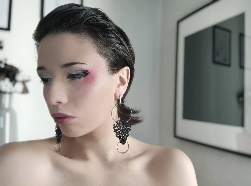  The image captures a poised model adorned with Myril Jewels' black and silver filigree hoop earrings, exuding an aura of neo-goth elegance. The intricate design of the earrings, with their delicate silver filigree, complements the model's dramatic eye makeup and bold, black lipstick. These earrings are perfect for those seeking a gothic-chic look, adding a touch of dark sophistication to any outfit, be it for Halloween, a rave party, or as a distinctive piece of everyday wear.